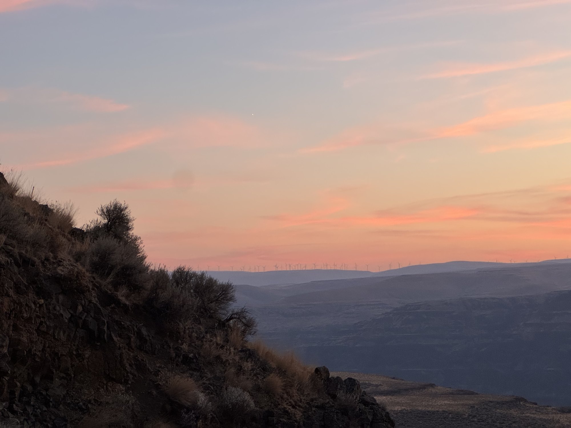 Sunset over a sagebrush plateau: a dark hillside in the foreground, hazy blue ridges beyond, and a distant line of wind turbines along the horizon under pink-streaked clouds.